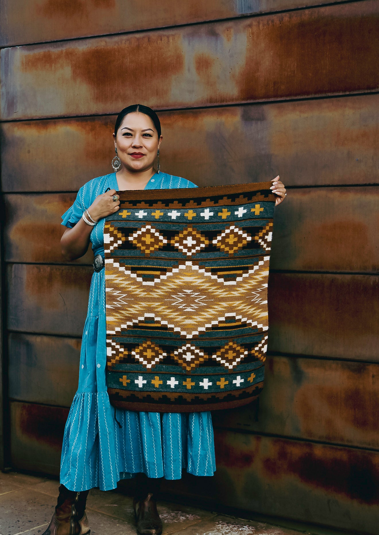 A woman stands in front of a rusted metal wall holding up a large woven textile with a Southwestern geometric pattern in brown, beige, black, and blue.